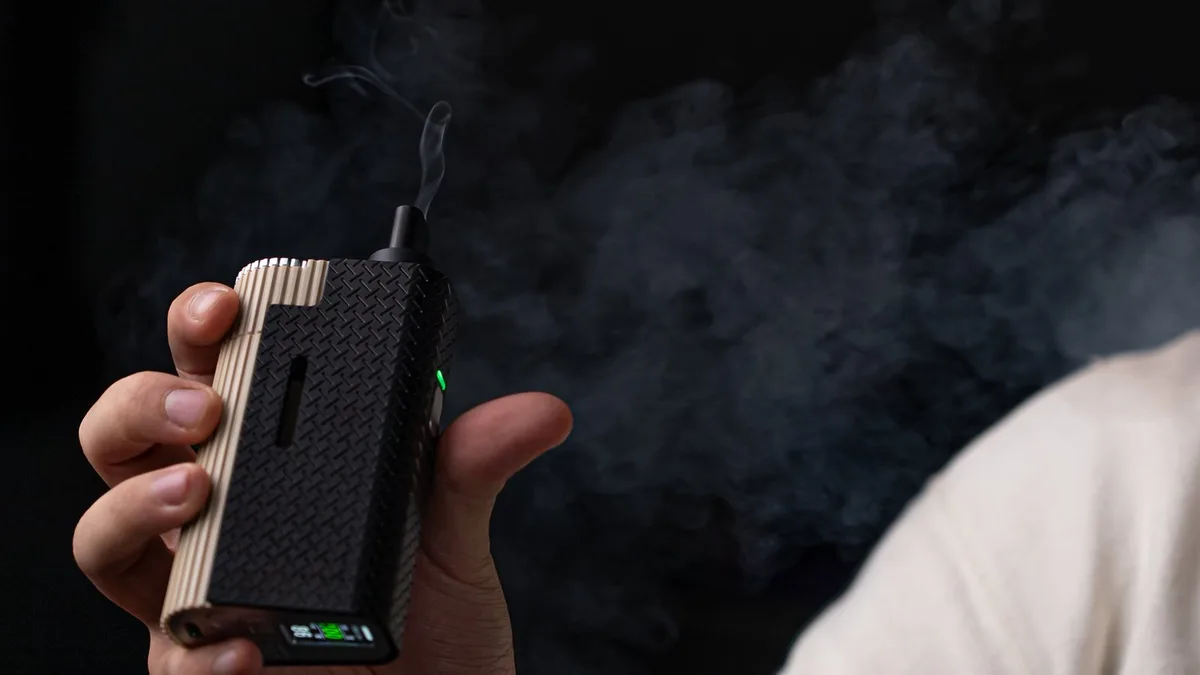 A hand holds a black and tan portable vaporizer with vapor rising from its mouthpiece against a dark, smoky background. | VapeExperts