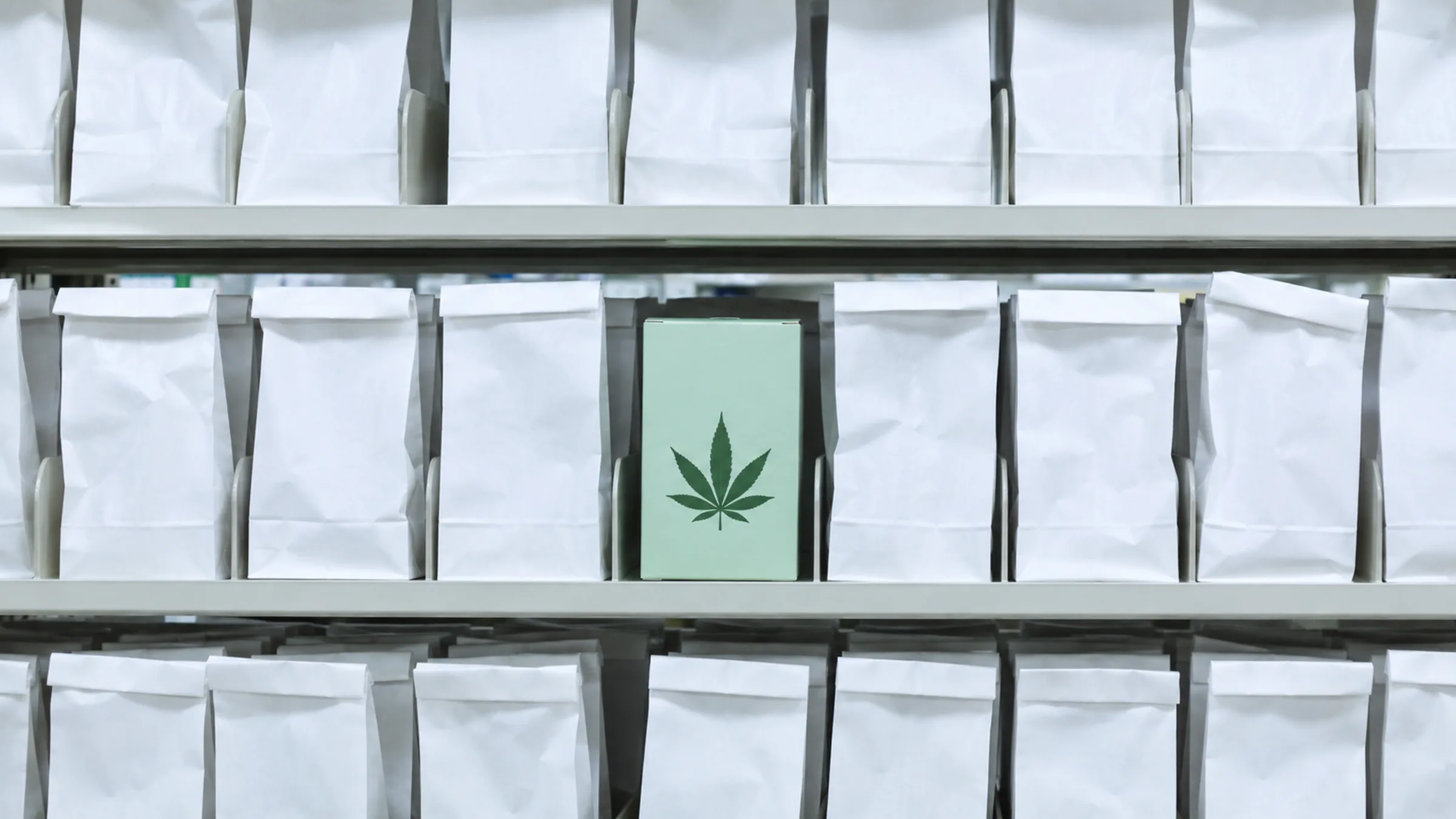 A pharmacy will-call shelf lined with rows of folded white prescription pickup bags, with one slot holding a mint-green medical cannabis box protruding slightly from the row. (VapeExperts/AI)