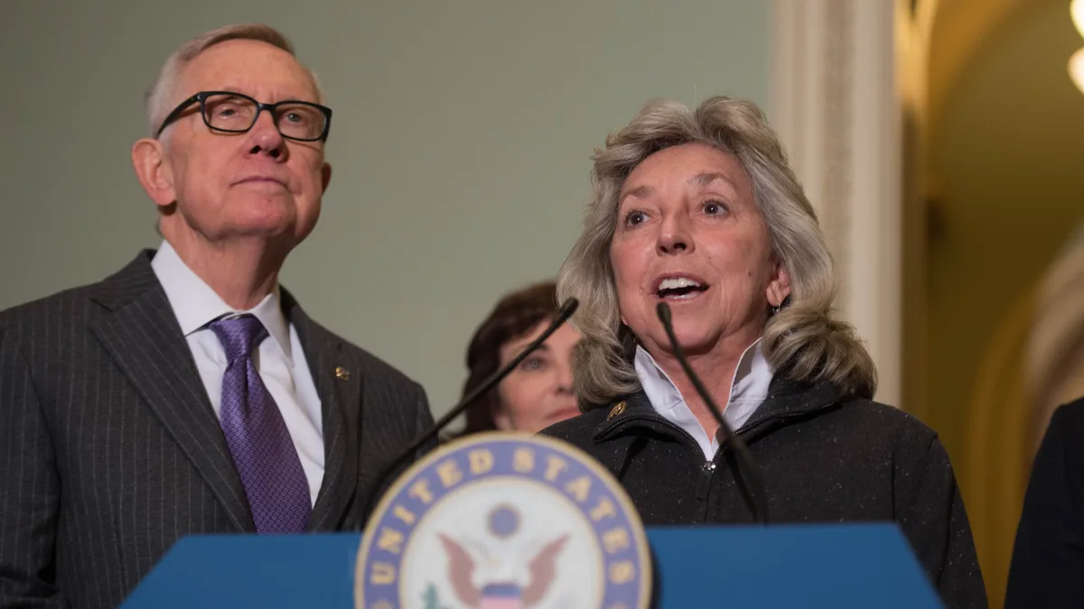 Two lawmakers at a blue podium with a United States Senate seal, a woman speaking into microphones while a man in glasses stands beside her. | Senate Democrats/Wikimedia Commons (CC BY 2.0)