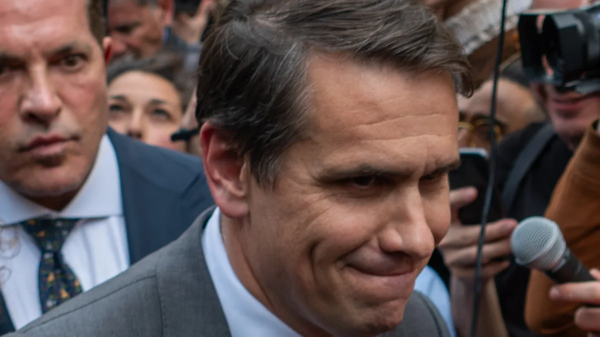 A man in a gray suit and red tie moves through a dense crowd of photographers and reporters holding cameras and microphones outside a courthouse. | BruceSchaff/Wikimedia Commons (CC BY-SA 4.0)