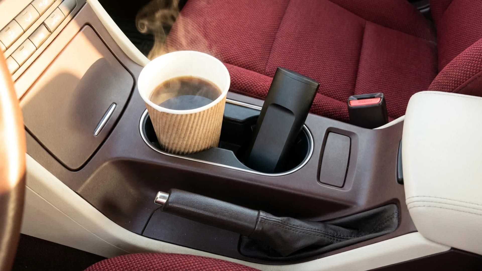 In der Mittelkonsole eines Autos steckt in einem Getränkehalter ein dampfender Kaffeebecher aus Papier und im anderen ein matter Vaporizer ohne Markenaufdruck für Cannabisblüten, daneben sind Gurtschloss und Wählhebel im warmen Morgenlicht zu sehen. (VapeExperts/AI)