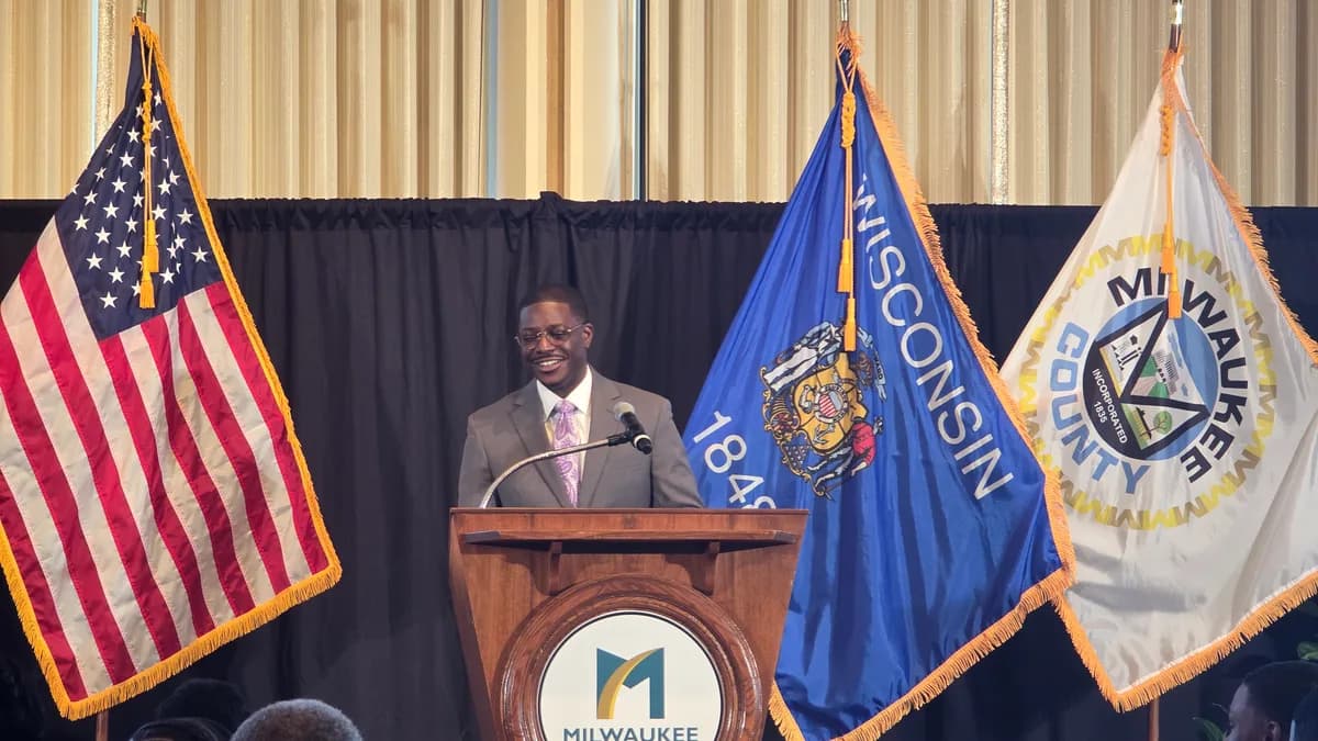 A man in a gray suit and purple tie smiles while speaking at a wooden podium bearing the Milwaukee County logo, with American, Wisconsin and... | HarvestLife/Wikimedia Commons (CC BY-SA 4.0)