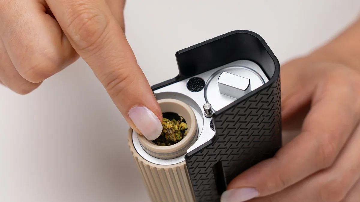 Close-up of hands holding a black and tan Frolic vaporizer while a finger presses ground herb into its open chamber. | VapeExperts