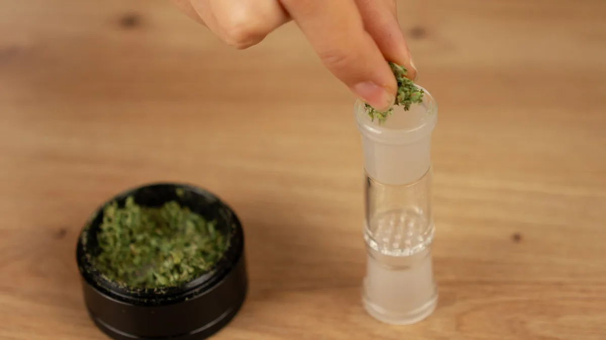 A hand places ground cannabis into a clear glass vaporizer bowl standing on a wooden table next to an open black grinder filled with herb. | VapeExperts