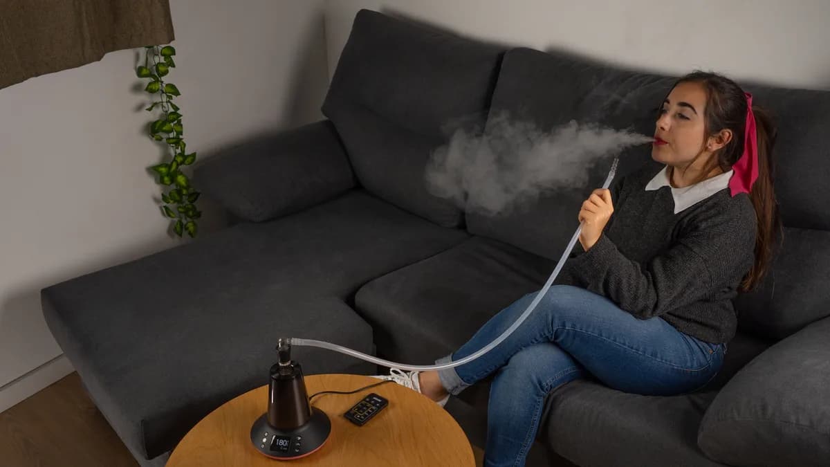 A woman sits on a dark couch exhaling vapor while holding a clear tube connected to an Arizer XQ2 vaporizer on a round wooden table. | VapeExperts