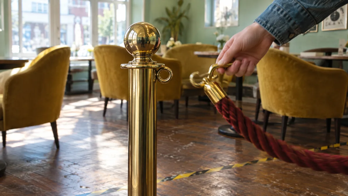 Tight crop of a brass floor stanchion and rope marking the edge of a lounge seating area, low armchairs beyond, taped boundary line on the floor.