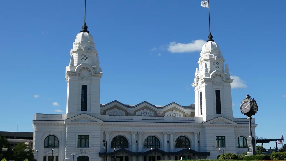 The white twin-towered Worcester Union Station under a blue sky, with an ornate street clock and hedges in front. | Pi.1415926535/Wikimedia Commons (CC BY-SA 4.0)