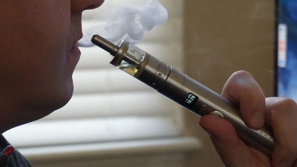 Close-up of a man exhaling vapor while holding a metal vape pen with a clear tank and digital display reading 13.2W to his lips. | Vaper City/Wikimedia Commons (CC BY-SA 4.0)