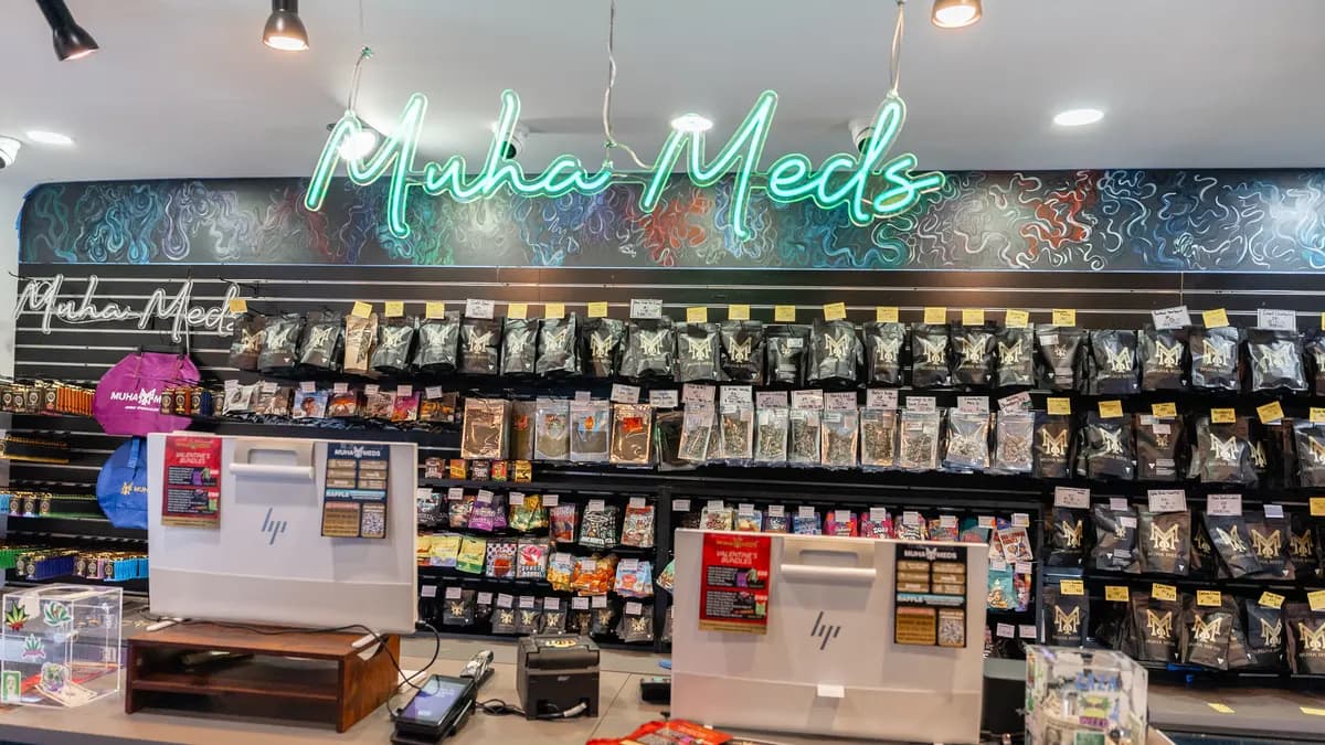 Interior of the Muha Meds Ypsilanti retail store with a green neon Muha Meds sign over walls of black branded cannabis bags and two register screens...