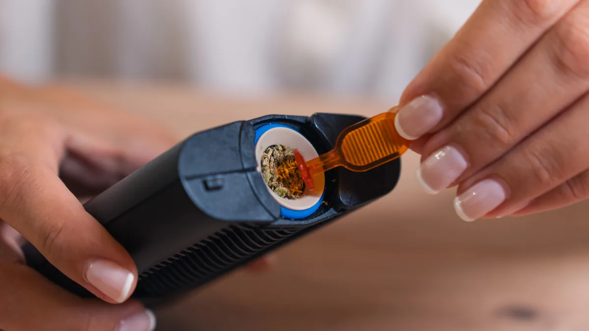 Close-up of hands using an orange tool to pack ground cannabis into the open chamber of a black Mighty+ vaporizer over a wooden table. | VapeExperts