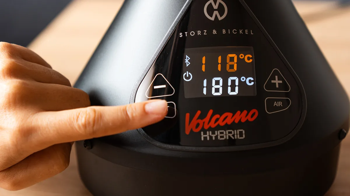 A finger presses a button on the control panel of a black Storz & Bickel Volcano Hybrid vaporizer sitting on a wooden table. | VapeExperts