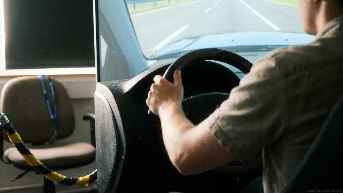 Research driving simulator: car seat and steering wheel rig facing wraparound curved screens showing a rendered highway, dim lab room, cables on floor.