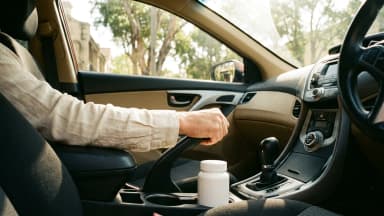 A white pharmacy medicine pot sits in a car's cupholder beside a hand holding the handbrake up, with sunlit gum trees blurred through the windscreen. (VapeExperts/AI)