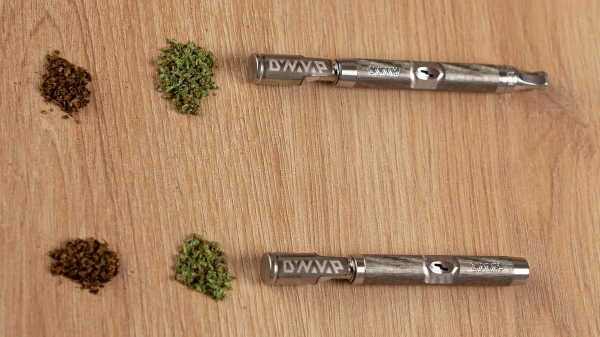 Two DynaVap M7 vaporizers lie on a wooden table next to piles of green ground cannabis and brown already-vaped bud. | VapeExperts